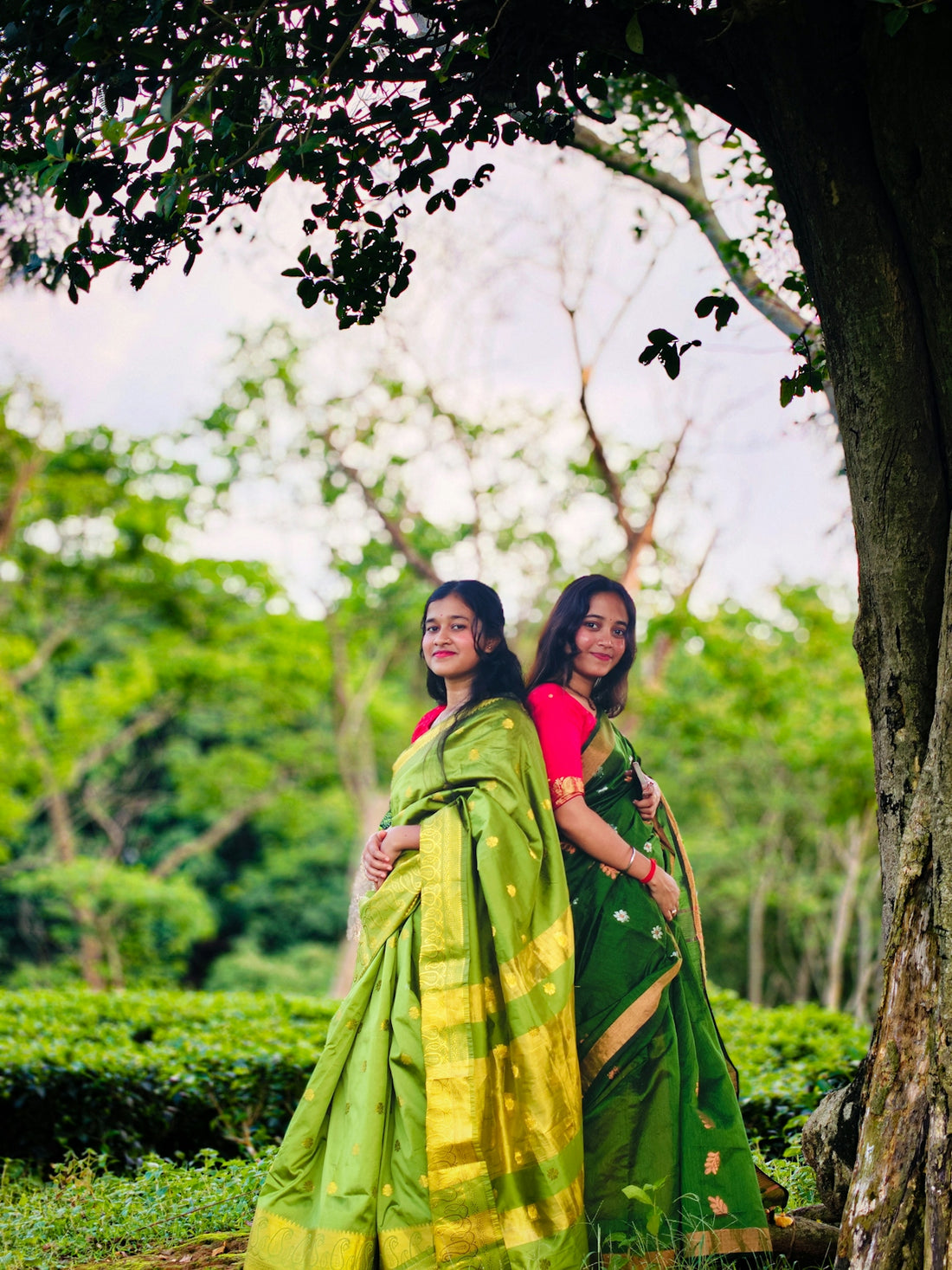 Two women pose in green saris by a tree.